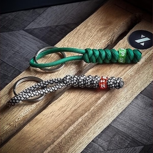 Puede incluir: Dos llaveros hechos a mano, uno verde y otro blanco y negro, descansan sobre una superficie de madera. Ambos tienen anillos metálicos y nudos decorativos. El llavero verde tiene cuentas verdes y blancas con letras. El llavero blanco y negro tiene cuentas rojas y blancas con letras.