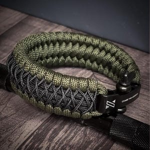 May include: A green and gray paracord bracelet with a black metal clasp. The bracelet is made of woven nylon cord and features the text "CHASE THE HORIZON" on the clasp. The bracelet is shown on a wooden surface.