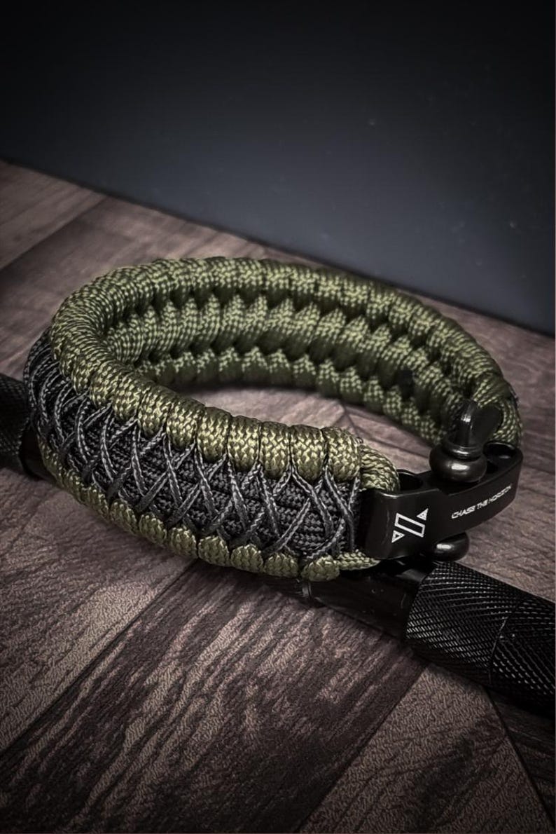 May include: A green and gray paracord bracelet with a black metal clasp. The bracelet is made of woven nylon cord and features the text "CHASE THE HORIZON" on the clasp. The bracelet is shown on a wooden surface.