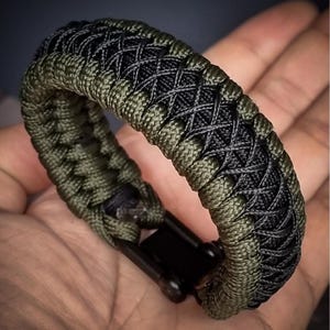 May include: A braided paracord bracelet in olive green and black, featuring a woven design. The bracelet has a black clasp and is shown on a hand.