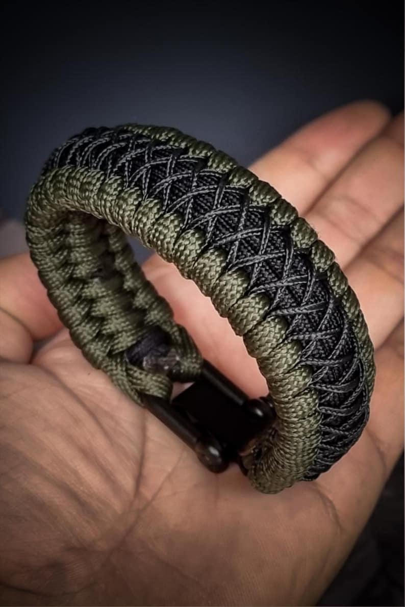 May include: A braided paracord bracelet in olive green and black, featuring a woven design. The bracelet has a black clasp and is shown on a hand.