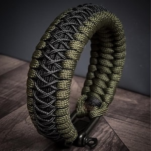 May include: A braided bracelet in olive green and black. The bracelet is made of woven cord, with a detailed pattern. The bracelet is shown on a dark wooden surface. The bracelet has a black metal clasp.