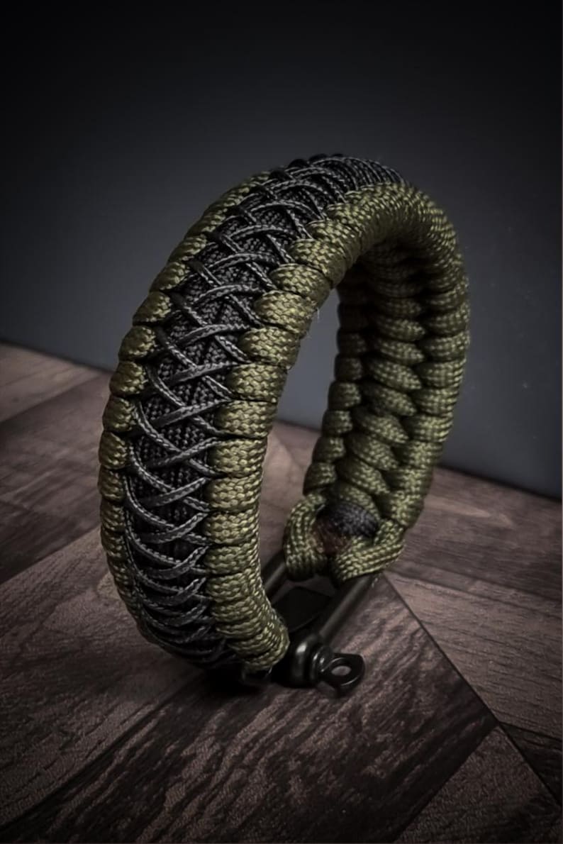 May include: A braided bracelet in olive green and black. The bracelet is made of woven cord, with a detailed pattern. The bracelet is shown on a dark wooden surface. The bracelet has a black metal clasp.