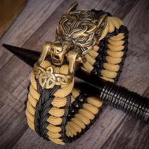 May include: A black and tan paracord bracelet with a wolf head clasp. The clasp is made of metal and has intricate details. The bracelet is a popular style for those who enjoy outdoor activities.