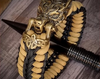 Puede incluir: Un brazalete de paracord negro y marrón con un cierre de cabeza de lobo. El cierre es de metal y tiene detalles intrincados. El brazalete es un estilo popular para quienes disfrutan de las actividades al aire libre.