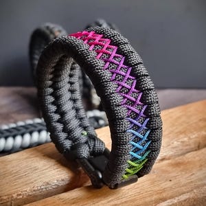 May include: Black paracord bracelet with a rainbow colored stitching pattern. The bracelet has a black metal clasp.