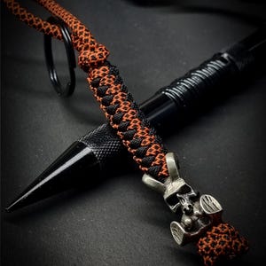 May include: A black tactical pen with an orange and black braided lanyard and a skull charm. The pen has a pointed tip and a textured grip. The lanyard is attached to a black keyring.