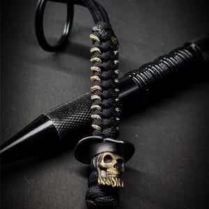 Op de afbeelding: Zwart paracord koord met een messing schedel kraal met een afstudeerhoed. Het koord is bevestigd aan een zwarte metalen ring. Een zwarte pen staat op de achtergrond. De schedel kraal heeft een verweerde afwerking.