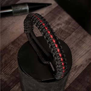 Può includere: Braccialetto intrecciato nero e rosso. Il braccialetto presenta un design intrecciato nero con una sottile striscia rossa al centro. Il braccialetto è realizzato in un materiale resistente ed è progettato per essere indossato al polso. È un accessorio elegante.