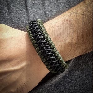 May include: A braided bracelet in shades of olive green and black. The bracelet is made of woven cord, with a textured appearance. The design features a repeating pattern of interwoven strands. The bracelet is worn on a wrist, against a gray background.
