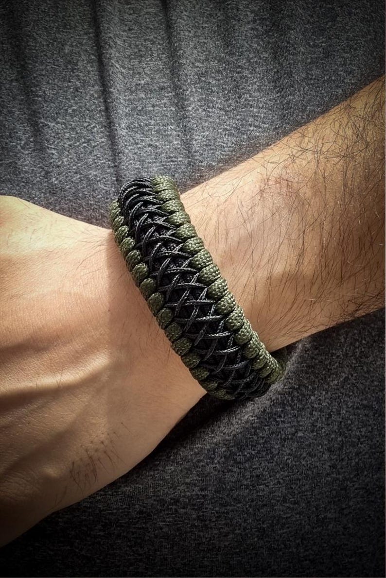 May include: A braided bracelet in shades of olive green and black. The bracelet is made of woven cord, with a textured appearance. The design features a repeating pattern of interwoven strands. The bracelet is worn on a wrist, against a gray background.