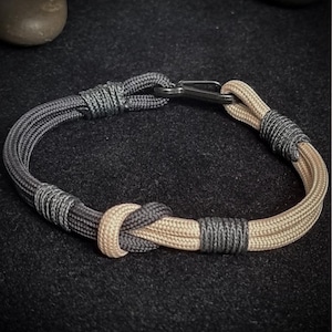 Puede incluir: Una pulsera de dos tonos con una combinación de cordón tejido negro y beige. La pulsera se asegura con un cierre de metal negro. El diseño incorpora nudos y secciones envueltas, creando un accesorio texturizado y elegante.
