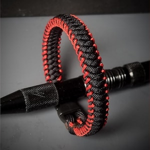 May include: Black and red woven bracelet with a black clasp. The bracelet is made of a tightly woven black cord with red accents. The bracelet is resting on a black object.