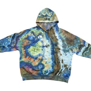 Op de afbeelding: Een tie-dye hoodie in tinten blauw, bruin, groen en oranje. De hoodie heeft een trekkoord en een kangoeroezak. Het verfpatroon creëert een gemarmerd effect over de stof. De hoodie ligt plat tegen een witte achtergrond.
