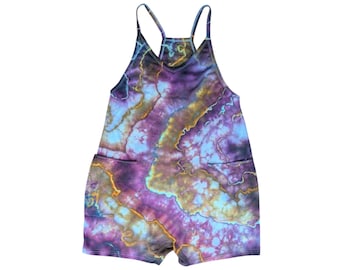 May include: A tie-dye romper in shades of purple, blue, and gold. The romper features a racerback design with thin straps, two front pockets, and a short leg. The fabric has a marbled effect.