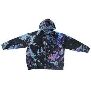 Op de afbeelding: Een zip-up hoodie met een tie-dye patroon in zwart, blauw en paars. De hoodie heeft een capuchon met trekkoord en geribde manchetten. Het tie-dye ontwerp is gewerveld en gemarmerd, wat een unieke uitstraling creëert.
