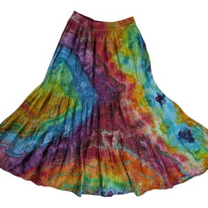 Op de afbeelding: Een levendige tie-dye rok in een vloeiend, gelaagd ontwerp. De rok heeft een spectrum aan kleuren, waaronder tinten blauw, groen, geel, oranje en paars. De stof lijkt lichtgewicht en geschikt voor warm weer.