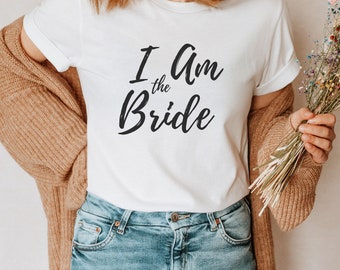 Puede incluir: Una camiseta blanca con el texto "I am the Bride" en letras cursivas negras. La camiseta la lleva puesta una persona que lleva pantalones vaqueros azules y un cárdigan marrón.