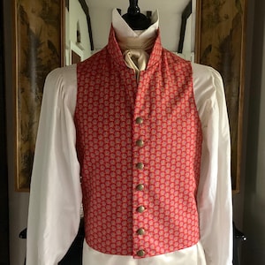 May include: A red patterned waistcoat with gold buttons. The waistcoat features a high collar and is worn over a white shirt. The pattern is a repeating floral design. The garment is part of a historical costume.