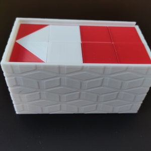 Può includere: Una scatola bianca con un motivo geometrico sul fondo e un puzzle rosso e bianco sulla parte superiore. Il puzzle è composto da piccoli quadrati e ha una forma triangolare al centro.