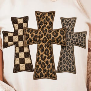 Könnte beinhalten: Ein cremefarbenes Kurzarm-T-Shirt mit einer Grafik aus drei kreuzförmigen Motiven im Stepp-Stil, die horizontal angeordnet sind. Das linke Kreuz hat ein braun-cremefarbenes Schachbrettmuster, das mittlere Kreuz ein braunes Leopardenmuster und das rechte Kreuz ein graues Leopardenmuster.
