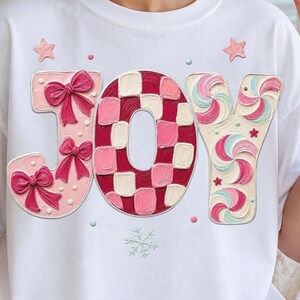 Peut inclure: Un t-shirt blanc avec le mot JOY écrit en grandes lettres stylisées. Le J est rose clair avec des nœuds rose foncé, le O présente un motif à carreaux rouges et roses, et le Y est orné de tourbillons de menthe rose et blanc avec des étoiles.