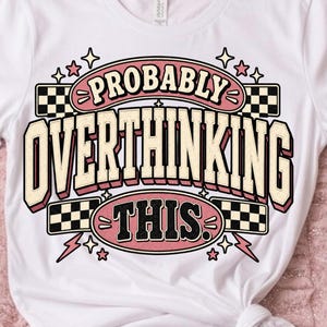 May include: A white short-sleeved t-shirt featuring a graphic design with the text "PROBABLY OVERTHINKING THIS" in bold lettering. The design includes black and white checkered patterns, pink accents, and small star and lightning bolt shapes.