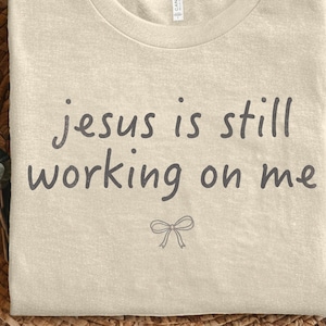 Könnte beinhalten: Ein hellbeiges Kurzarm-T-Shirt mit dem dunkelgrauen Schriftzug "jesus is still working on me" in einer einfachen, abgerundeten Schriftart auf der Brust und einem kleinen, minimalistischen Strichzeichnungs-Schleifensymbol unter dem Text.