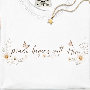 Könnte beinhalten: Ein weißes T-Shirt mit einem Comfort Colors-Etikett in Größe XL. Das Shirt zeigt einen braunen Schriftzug "peace begins with Him", umgeben von Illustrationen von Gänseblümchen und zwei kleinen Schmetterlingen. Eine goldene Sternkette liegt darauf.