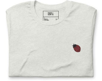 May include: A light gray t-shirt with a small embroidered ladybug on the front. The shirt has a crew neck and a tag that reads "FOURTH & GLORY CO." and "Made in Nicaragua."