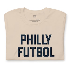 May include: A folded, cream-colored t-shirt with the words "PHILLY FUTBOL" printed in navy blue block letters. The shirt has a crew neck and short sleeves. The tag reads "Made in Nicaragua" and "100% combed ring spun cotton".