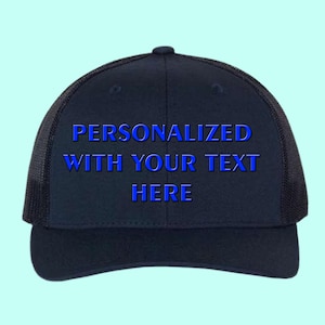 May include: A navy blue trucker hat with a mesh back and a curved brim. The front of the hat features the text "PERSONALIZED WITH YOUR TEXT HERE" in blue, indicating a customizable design. The hat is set against a light blue background.