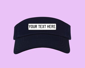 May include: Navy blue visor with a white rectangular patch that reads "YOUR TEXT HERE". The visor has a curved brim and is set against a light purple background. This hat is suitable for sports or outdoor activities.