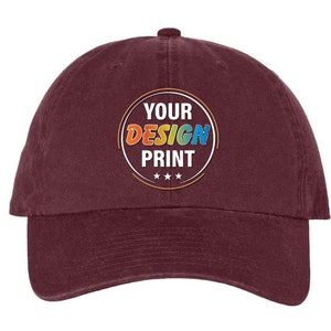 May include: A maroon baseball cap with a white circle and the text "YOUR DESIGN PRINT" in colorful letters.