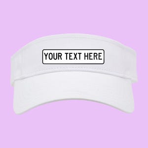 May include: A white visor with a curved brim. The visor has a rectangular black and white label that reads "YOUR TEXT HERE". The background is a solid light purple color.