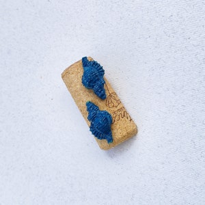 May include: Two blue seashell-shaped earrings on a cork background. The cork has the word "Bottles" printed on it.