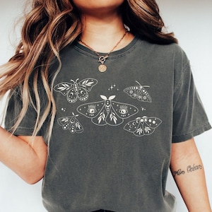 May include: A dark gray t-shirt with a white graphic of several moths with celestial details. The moths have eyes, moons, and stars on their wings.