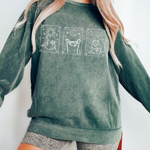Puede incluir: Una sudadera verde oscuro con un gr&aacute;fico blanco de tres ilustraciones de gatos en marcos rectangulares. Las ilustraciones de gatos representan el sol, la luna y las nubes. La sudadera se usa con pantalones cortos grises.