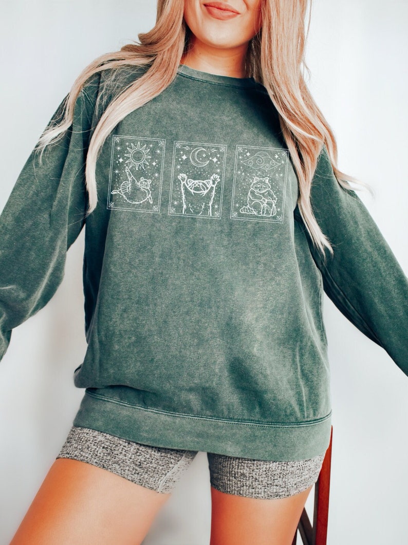 Puede incluir: Una sudadera verde oscuro con un gr&aacute;fico blanco de tres ilustraciones de gatos en marcos rectangulares. Las ilustraciones de gatos representan el sol, la luna y las nubes. La sudadera se usa con pantalones cortos grises.