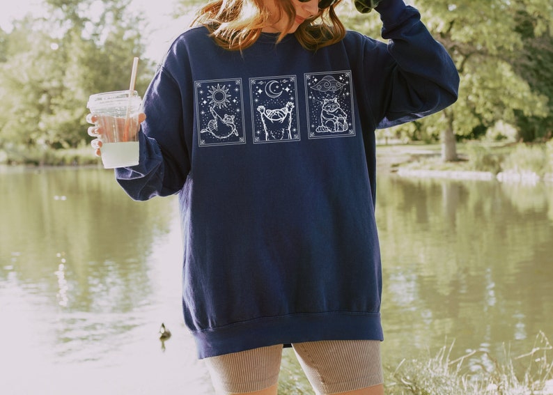 Puede incluir: Sudadera azul marino con tres paneles ilustrados en blanco. Cada panel presenta un gato con elementos celestiales: sol, luna y nubes. La sudadera tiene un corte holgado, perfecta para uso casual.