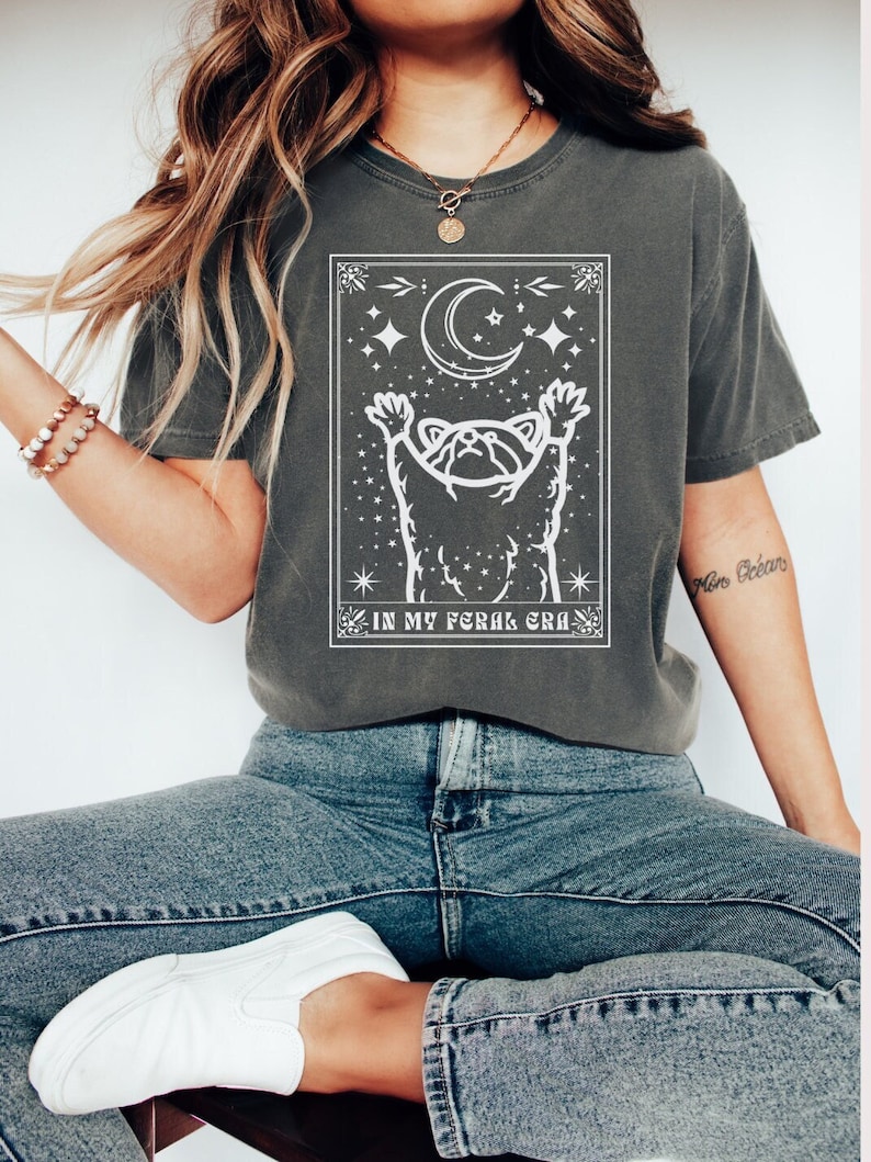 Puede incluir: Una camiseta gris oscuro con un gr&aacute;fico blanco de un mapache que alcanza una luna creciente con estrellas. El texto "IN MY FERAL ERA" est&aacute; debajo del mapache.