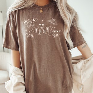 May include: A brown oversized t-shirt with a white graphic of moths and butterflies. The moths have eyes and crescent moons on their wings.