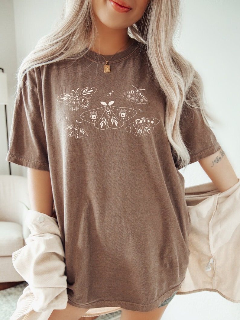 May include: A brown oversized t-shirt with a white graphic of moths and butterflies. The moths have eyes and crescent moons on their wings.