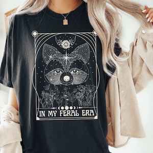 Puede incluir: Camiseta negra con un dise&ntilde;o gr&aacute;fico blanco de la cara de un animal rodeado de flores y el texto "IN MY FERAL ERA".