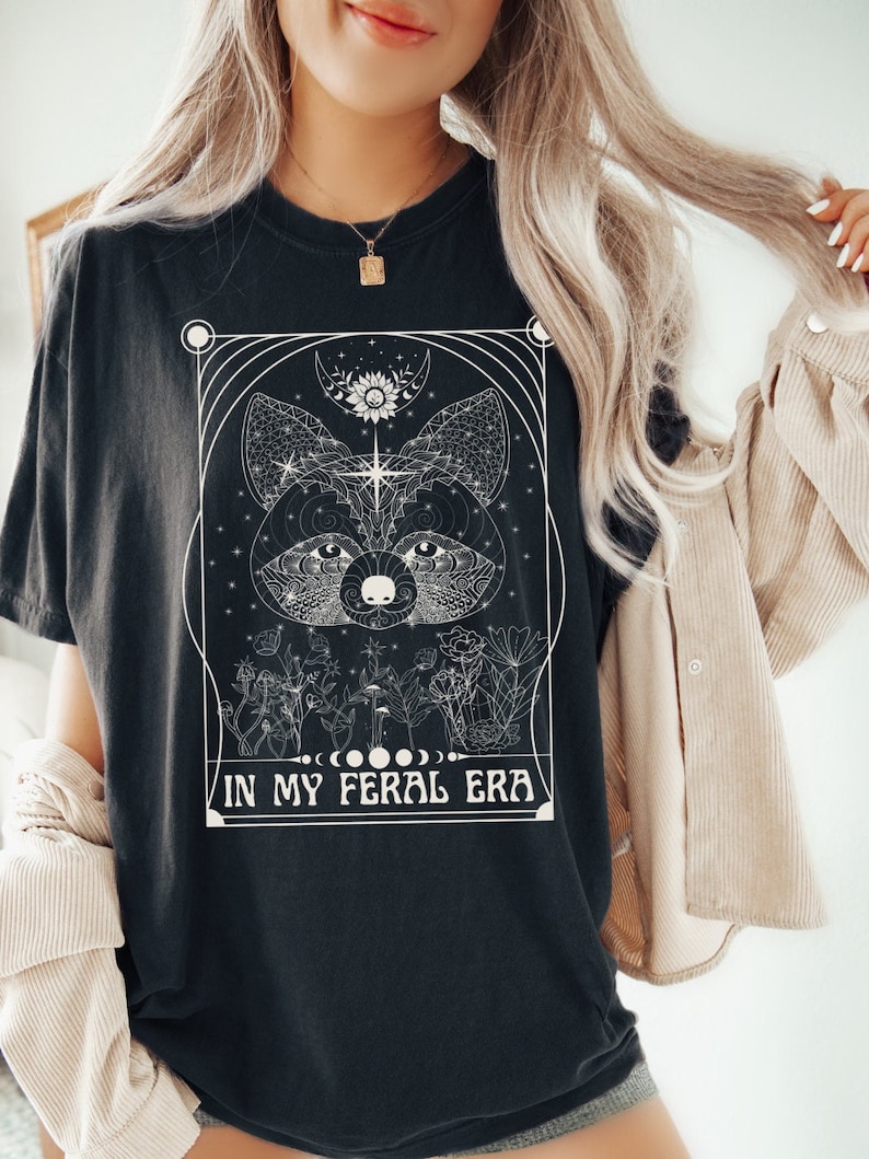Puede incluir: Camiseta negra con un dise&ntilde;o gr&aacute;fico blanco de la cara de un animal rodeado de flores y el texto "IN MY FERAL ERA".