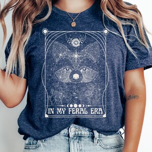 Puede incluir: Una camiseta azul marino con un dise&ntilde;o gr&aacute;fico blanco de la cara de un mapache rodeado de formas geom&eacute;tricas y el texto "IN MY FERAL ERA".