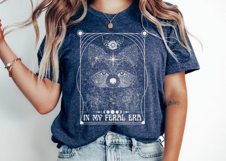 Puede incluir: Una camiseta azul marino con un dise&ntilde;o gr&aacute;fico blanco de la cara de un mapache rodeado de formas geom&eacute;tricas y el texto "IN MY FERAL ERA".