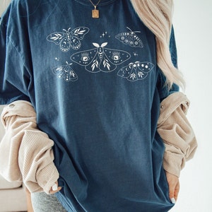 May include: A dark blue oversized t-shirt with a white graphic of moths and celestial elements. The moths have intricate details and are surrounded by stars and moons.