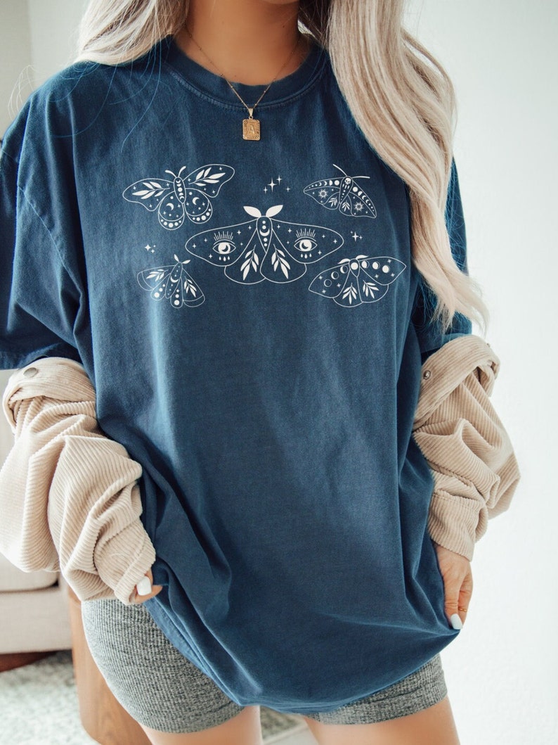 May include: A dark blue oversized t-shirt with a white graphic of moths and celestial elements. The moths have intricate details and are surrounded by stars and moons.