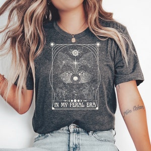 Puede incluir: Una camiseta gris oscuro con un gr&aacute;fico blanco de la cara de un mapache rodeado de estrellas y cuerpos celestes. El texto "IN MY FERAL ERA" est&aacute; impreso debajo del gr&aacute;fico.
